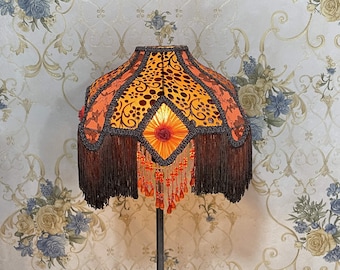 Op de afbeelding: Een lampenkap met een franje rand en kralen accenten. De kap is gemaakt van stof met een bloemenpatroon in tinten oranje, geel en bruin.
