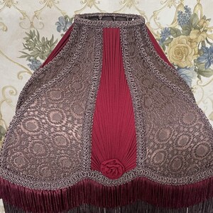 Puede incluir: Una pantalla de l&aacute;mpara roja y marr&oacute;n con un dise&ntilde;o plisado y un ribete de flecos. La pantalla tiene un patr&oacute;n floral y un borde festoneado.