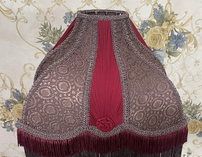 Puede incluir: Una pantalla de l&aacute;mpara roja y marr&oacute;n con un dise&ntilde;o plisado y un ribete de flecos. La pantalla tiene un patr&oacute;n floral y un borde festoneado.