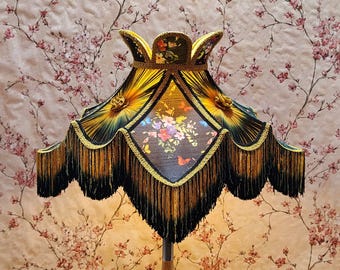 May include: A decorative lampshade with a black, gold, and yellow color scheme. The lampshade features a floral and butterfly design, with a fringe trim. The lampshade is a vintage style and is likely used for home decor.