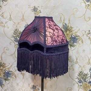 K&ouml;nnte beinhalten: Ein lila und schwarzer Lampenschirm mit Fransenbesatz. Der Lampenschirm ist aus Stoff und Spitze gefertigt und hat ein florales Design.