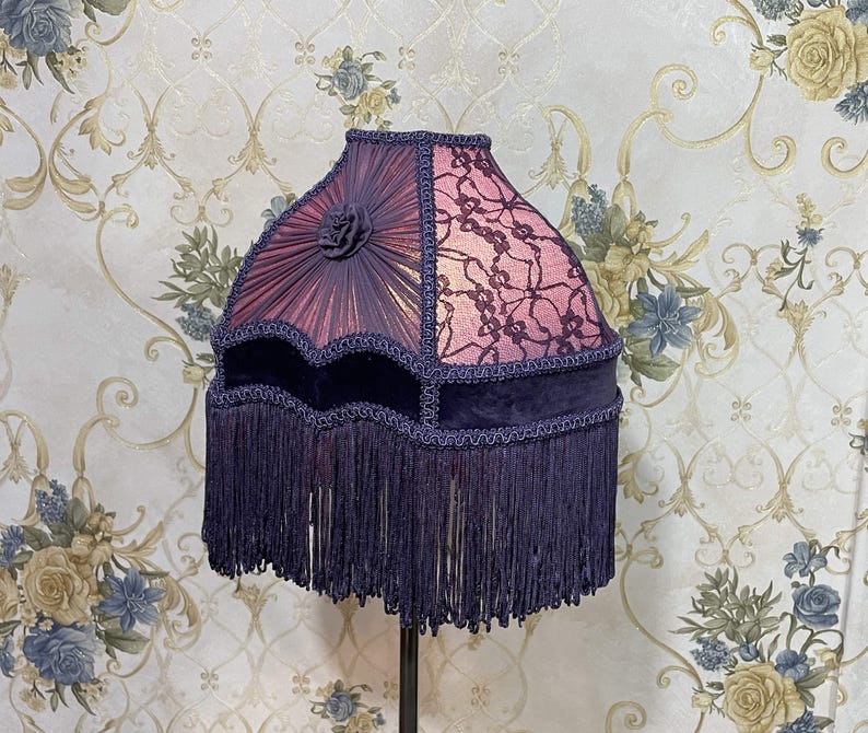 K&ouml;nnte beinhalten: Ein lila und schwarzer Lampenschirm mit Fransenbesatz. Der Lampenschirm ist aus Stoff und Spitze gefertigt und hat ein florales Design.