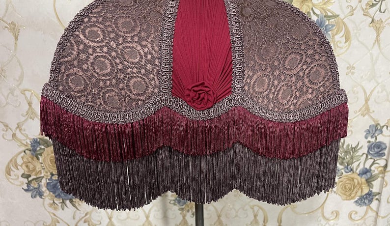 Puede incluir: Una pantalla de l&aacute;mpara color burdeos con flecos festoneados y una parte superior de tela estampada. La tela tiene un delicado dise&ntilde;o floral y un ribete burdeos.
