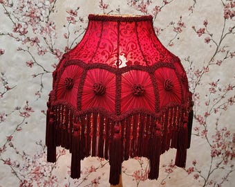 Könnte beinhalten: Roter Lampenschirm mit floralem Muster und dekorativen Fransen. Der Lampenschirm hat ein detailliertes Design mit Rosetten und Quasten. Ein dekorativer Einrichtungsgegenstand.