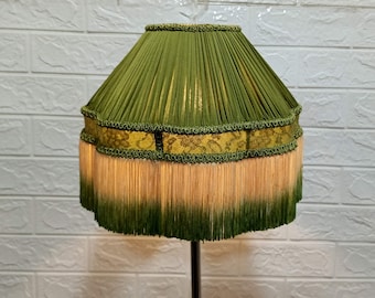 Può includere: Un paralume verde con bordatura dorata e una frangia di nappe verdi e beige.