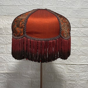 May include: A red lampshade with a gold and brown patterned fabric and a fringe trim.
