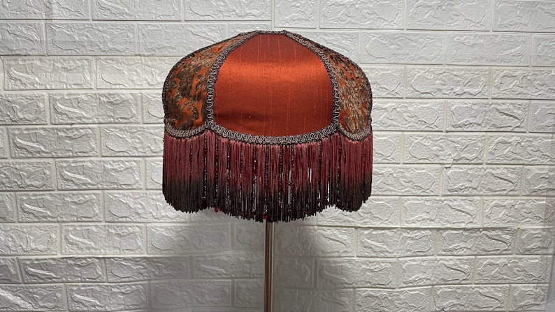 May include: A red lampshade with a gold and brown patterned fabric and a fringe trim.