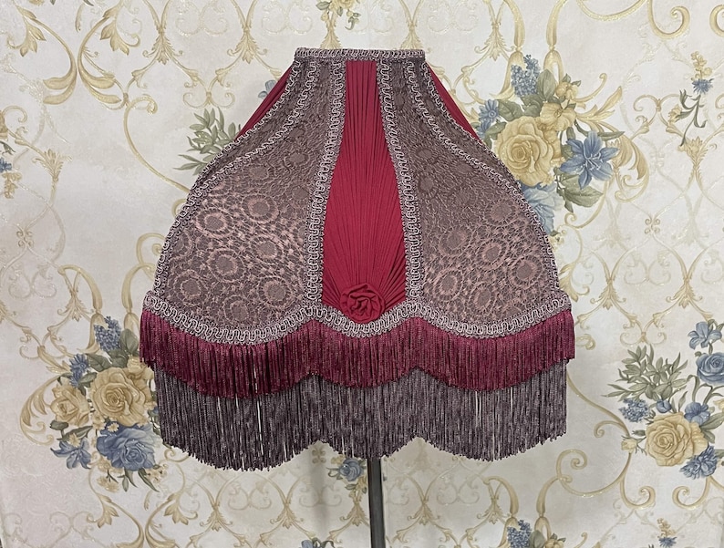 Puede incluir: Una pantalla de l&aacute;mpara de color burdeos con un centro de tela plisada y un ribete de encaje decorativo. La pantalla tiene una doble capa de flecos burdeos.