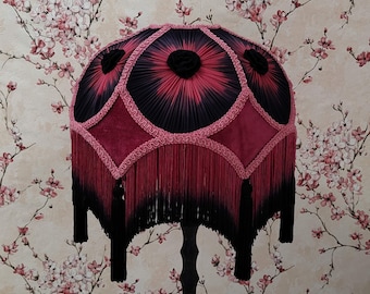 Può includere: Paralume decorativo nero e rosso, con un design a pieghe e un pannello a forma di diamante. Ha frange ed è posto su una base nera. Lo sfondo è una carta da parati floreale. Un oggetto decorativo.