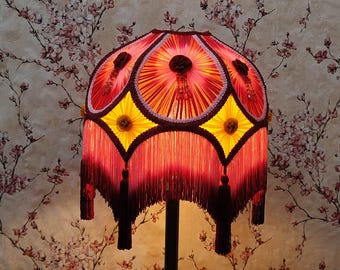 May include: A decorative lampshade with a vibrant, layered design. The shade features a red and yellow color scheme with intricate patterns, tassels, and a fringed bottom. The lamp is supported by a black stand.