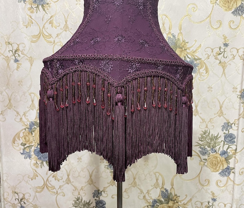 K&ouml;nnte beinhalten: Ein lilafarbener Lampenschirm mit Fransenbesatz und Perlenakzenten. Der Lampenschirm ist aus einem Stoff mit Blumenmuster.