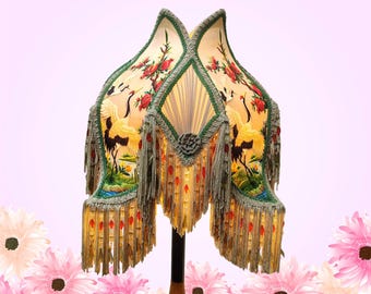 May include: A multi-paneled fabric lampshade featuring a cream background with printed illustrations of cranes, red flowers, and green foliage. The shade is adorned with green braided trim, long gray fringe, and red bead accents.