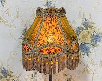May include: A gold and orange lampshade with a beaded fringe and a floral pattern. The lampshade is made of fabric and has a scalloped edge.