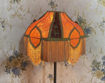 May include: A vintage-style lampshade with a geometric pattern in shades of orange and brown. The shade is trimmed with gold fringe and beads.