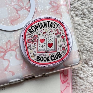 以下が含まれることがあります： ピンクと白の携帯電話ケースに、白いリングホルダーと、ピンクと白の「Romanstasy Book Club」と書かれたステッカーが付いています。ステッカーには、本、ハート、星が描かれています。