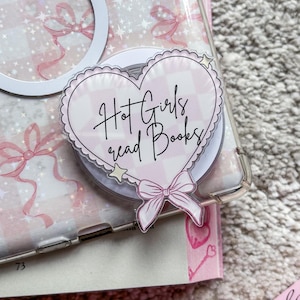 May include: A pink and white heart-shaped sticker with the text "Hot Girls read Books" in black cursive writing. The sticker has a white bow and a small white star on it. The sticker is on a pink and white patterned phone case.