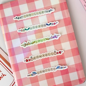 Puede incluir: Un cuaderno a cuadros rosas y blancos con cinco pegatinas que dicen "THRILLER", "HORROR", "SMUT", "ROMANCE" y "FANTASY". Un libro rosa con las palabras "Bad Craz" está en el fondo. Una tarjeta que dice "Please to the beach" también está presente.