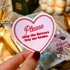 Op de afbeelding: Een hartvormige sticker met een roze achtergrond en een rode geschulpte rand. De sticker leest "Please skip the flowers buy me books" in rode tekst. De sticker wordt in een hand gehouden.