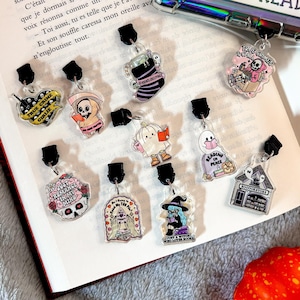 Peut inclure: Une collection de 10 breloques en acrylique transparent avec différents motifs sur le thème d'Halloween. Les breloques présentent des illustrations d'une sorcière lisant un livre, un crâne avec un cerveau, un fantôme lisant un livre, une librairie, une citrouille, un squelette, un livre avec un cœur, un fantôme, un chaudron et un livre avec un crâne. Les breloques sont attachées à des anneaux en métal noir.