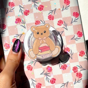 Pode incluir: Uma capa transparente para telefone com um padrão xadrez rosa e branco e um design de rosas cor-de-rosa. Um urso de peluche castanho com um laço rosa e uma pilha de livros está preso à capa com um anel transparente.