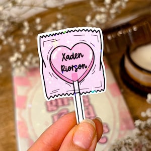 Peut inclure: Une sucette rose en forme de cœur avec le texte "Xaden Riotson" dessus. La sucette est enveloppée dans un papier rose et blanc avec des accents de paillettes. La sucette est sur un bâton.