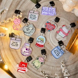 Peut inclure: Une collection de 10 charmes de livre colorés présentant divers motifs, notamment un crâne, une grenouille, un cœur, un livre, un globe à neige, un dé et un bocal. Les charmes sont en plastique transparent avec des détails et du texte colorés. Le texte sur les charmes comprend des phrases comme "Smells like she's reading Fantasy again", "Emotional Support Kindle", et "Pretty Girls Read".