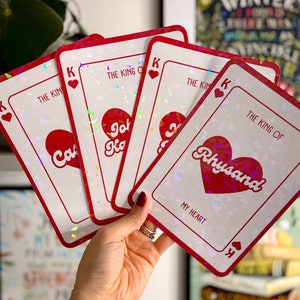 以下が含まれることがあります： 赤いハートのデザインと「The King of」というテキストと名前が書かれた4枚のトランプ。カードは虹色で、キラキラした仕上げになっています。