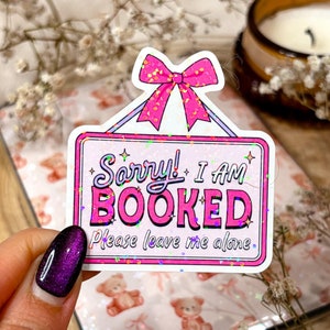 May include: Pink and white glitter sticker with a sign that says "Sorry! I am booked. Please leave me alone." The sign has a pink bow on top and is surrounded by glitter.