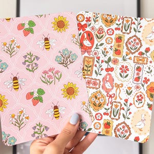Op de afbeelding: Twee decoratieve papieren vellen met bloemen- en insectenpatronen. Eén vel heeft een roze achtergrond met bijen, zonnebloemen en aardbeien. Het andere vel heeft een witte achtergrond met omlijste bloemen- en ganzenontwerpen. De vellen worden in een hand gehouden.