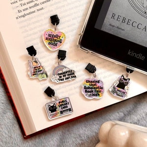 Peut inclure: Une collection de six breloques acryliques colorées avec différents motifs sur le thème du livre. Les breloques sont attachées à des clips noirs et comprennent des phrases comme "Silently Screaming Inside", "Currently in my Fuck This Shit Era", "Chasing that Scholastic Book Fair High", et "Books are Therapy".