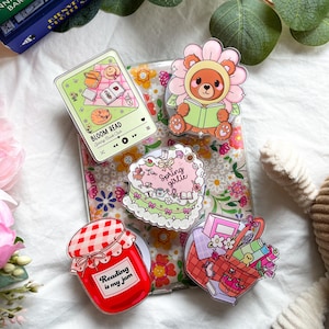 Puede incluir: Un conjunto de coloridos agarres acrílicos para teléfonos con varios diseños. Incluye un tarro de mermelada rojo con "Reading is my jam", un oso en una flor, una cesta de picnic, un pastel y un diseño "Bloom Read".