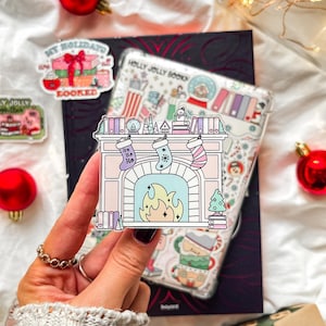 Könnte beinhalten: Eine transparente Handyhülle mit weihnachtlichem Design, die Strümpfe, einen Kamin und Bücher zeigt. Die Hülle wird über einem dunklen Buch gehalten, mit Aufklebern und roten Kugeln verstreut. Die Aufkleber lauten "My Holidays are Booked" und "Holly Jolly Bookin".