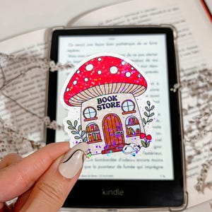 May include: A whimsical sticker featuring a mushroom-shaped bookstore with a red glittery cap and the text "BOOK STORE". The sticker has a brown door, windows, and a small cat. The sticker is held in front of a Kindle.