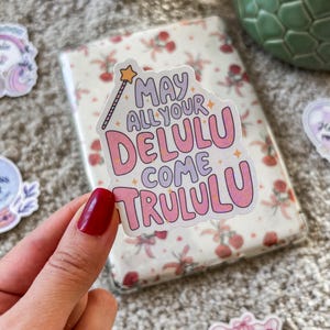 Peut inclure: Un autocollant rose et blanc avec un design fantaisiste et le texte "May all your Delulu come Trululu".