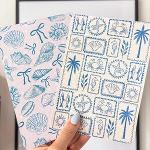May include: Two notebooks with seaside designs. One has a pale pink background with blue seashells and bows. The other has a cream background with blue stamps of palm trees, starfish, and seahorses. The notebooks are held by a person.