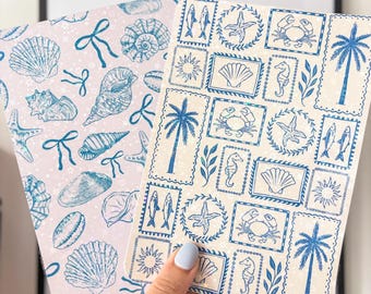 Könnte beinhalten: Zwei Notizbücher mit Strandmotiven. Eines hat einen hellrosa Hintergrund mit blauen Muscheln und Schleifen. Das andere hat einen cremefarbenen Hintergrund mit blauen Briefmarken von Palmen, Seesternen und Seepferdchen. Die Notizbücher werden von einer Person gehalten.