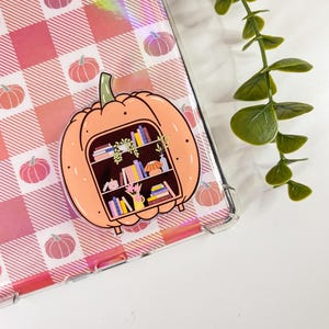 Könnte beinhalten: Ein orangefarbener Kürbis-Aufkleber mit einem Bücherregal-Design. Der Aufkleber hat einen dunkelbraunen Innenraum mit Miniaturbüchern, Pflanzen und einer Lampe. Der Hintergrund ist ein rosa-weißes Karomuster mit Kürbis-Illustrationen.