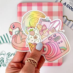 Op de afbeelding: Een verzameling kleurrijke stickers met zomerthema's. De stickers bevatten een roze flamingo-drijver, een ijsje, een camera, een zonnebril en een lolly. De stickers worden in een hand met een gouden ring gehouden, tegen een roze en witte geruite achtergrond.