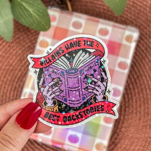 Peut inclure: Autocollant holographique avec le texte "Villains Have The Best Backstories". Il représente un livre violet tenu par des mains squelettiques avec des bijoux dorés. Le sticker a une bannière rouge.