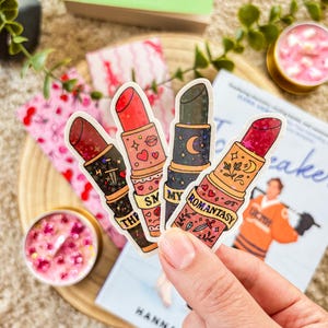 Könnte beinhalten: In einer Hand werden vier Lippenstift-förmige Aufkleber mit verschiedenen Designs und Farben gehalten. Die Aufkleber zeigen rote, rosa und dunkle Lippenstiftfarben mit dekorativen Elementen wie Herzen, Monden und floralen Mustern. Im Hintergrund sind Bücher und Kerzen zu sehen.