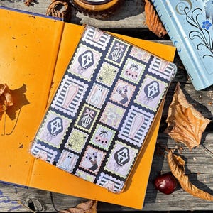May include: A clear phone case featuring a repeating pattern of Halloween-themed postage stamps. The stamps include images of ghosts, coffins, and the Scream mask. The case is set on an open orange book, with a candle and a blue book in the background.