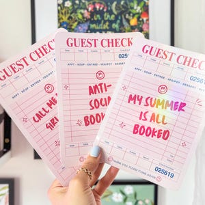 May include: Pink guest check pads with various phrases in colorful text, including "Call Me Siri," "Anti-Social Book," and "My Summer Is All Booked." Each pad has a check number and a smiley face.