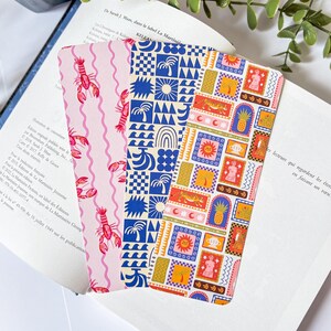Peut inclure: Trois marque-pages décoratifs aux motifs colorés sont présentés dans un livre ouvert. L'un des marque-pages a un fond rose avec des illustrations de homards rouges. Le deuxième marque-page a un motif géométrique bleu et blanc. Le troisième a un motif coloré.