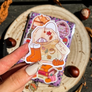 Könnte beinhalten: Ein Aufkleber mit einem Teebeutel-Design, verziert mit Herbstillustrationen. Er zeigt orangefarbene Gummistiefel, Kürbisse, eine Tasse Tee und den Text "Cozy Reader Tea". Der Hintergrund umfasst einen Stapel Bücher und einen lila Einband.
