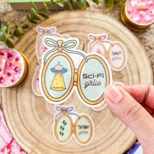 Peut inclure: Plusieurs autocollants avec un design de médaillon. L'un d'eux représente un OVNI et les mots "Sci-Fi girlie". D'autres autocollants disent "Shut girlie" et "Thriller girlie". Les autocollants sont sur une surface en bois.