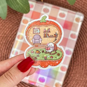 May include: A sticker featuring a pumpkin-shaped scene with a cozy interior. Inside, a small character reads in a rocking chair. The sticker includes the text "Cozy Reads" and is surrounded by a glittery border.