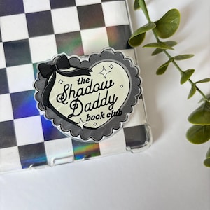 Op de afbeelding: Een hartvormige sticker met de tekst "the Shadow Daddy book club" in een zwart-wit ontwerp. De sticker heeft een geschulpte rand en een zwarte strik. De achtergrond is een zwart-wit geblokt patroon.