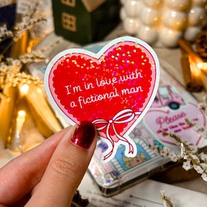 Peut inclure: Un autocollant en forme de cœur rouge avec des paillettes et une bordure blanche. L'autocollant a un ruban blanc et le texte "Je suis amoureuse d'un homme fictif".