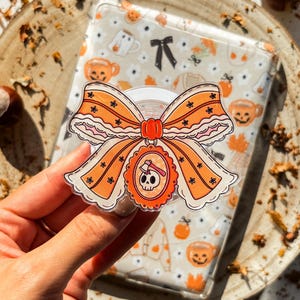 Puede incluir: Una funda transparente para teléfono con un diseño de Halloween, con calabazas naranjas, tazas de té y un lazo negro. Un lazo naranja decorativo con un dije de calavera y hacha está adjunto. La funda está sujeta por una mano.