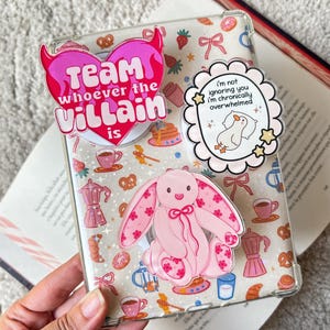Könnte beinhalten: Transparente Handyhülle mit rosa-weißem Muster, mit einem rosa Hasen und mehreren Aufklebern. Die Aufkleber enthalten ein herzförmiges mit dem Text "Team whoever the Villain is" und ein rundes mit einer Ente und dem Text "I'm not ignoring you I'm chronically overwhelmed."